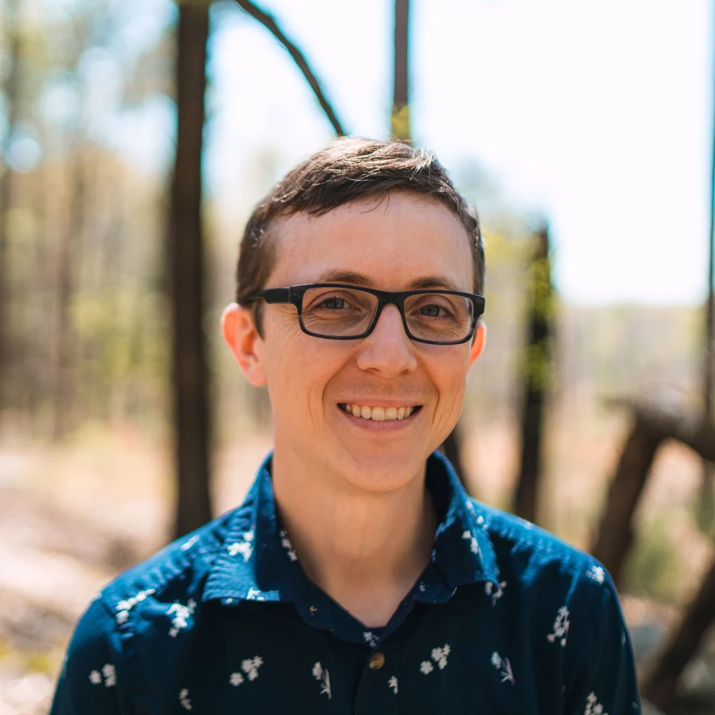 An outdoor portrait of Timothy wearing a dark blue shirt with a floral patter and black, plastic framed glasses.